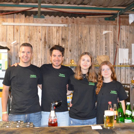 Vier Personen lächeln für ein Foto in einer Bar, tragen passende schwarze T-Shirts mit grünem Text. Dahinter sind hölzerne Wände und Regale mit Gegenständen wie Flaschen und Gläsern zu sehen. Im Vordergrund befindet sich eine Theke mit Gläsern und Flaschen.