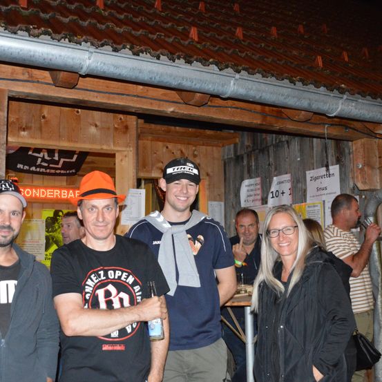 Eine Gruppe von Menschen, darunter ein Mann mit einer Flasche, steht nachts vor einer Bar. Dahinter hält ein Mann ein Glas, und eine Frau lächelt. Poster an der Wand werben für Zigaretten und Snus.