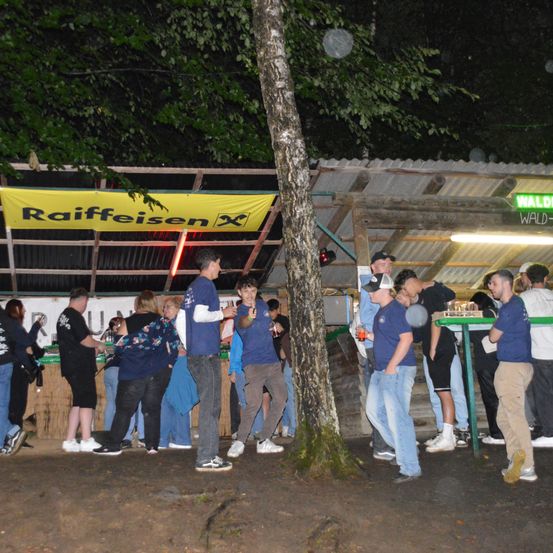 Eine Nachtszene mit Menschen, die um eine Bar stehen, beleuchtet von roten Lichtern, unter einem Banner mit dem Wort Raiffeisen.