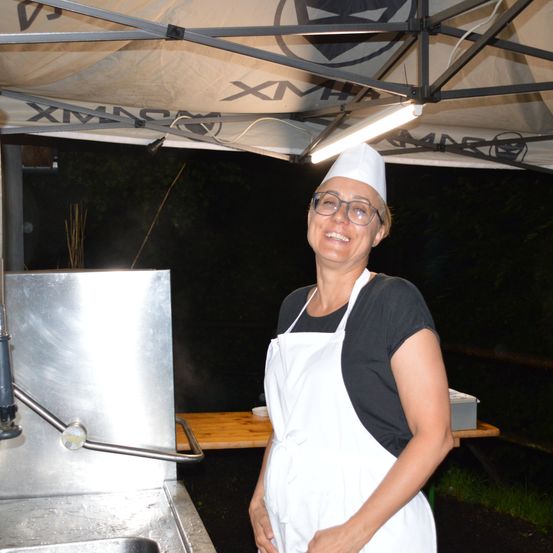 Eine lächelnde Frau in einer Kochmütze und einem weißen Schürze steht unter einem Baldachin und hält die Hände in die Hüften gestemmt. Sie befindet sich in der Nähe eines Edelstahlspülbeckens mit Wasserhahn und einem Holztisch.