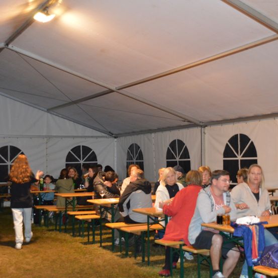 Eine Gruppe von Menschen sitzt an Tischen in einem großen Zelt, wahrscheinlich bei einer Zusammenkunft. Einige trinken und unterhalten sich. Eine Frau geht im Vordergrund.