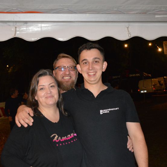 Drei Menschen lächeln und posieren für ein Foto unter einem Zelt bei Nacht. Der Mann auf der rechten Seite trägt ein schwarzes Shirt mit 'BURSCHENSCHAFT' und 'ANGECK' darauf geschrieben.