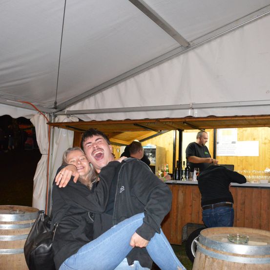 Zwei Menschen lachen und umarmen sich in einem Zelt mit Tresen und Holzfässern.