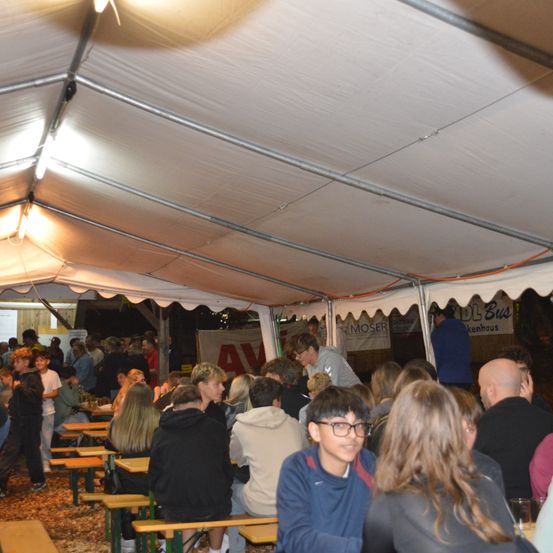Eine Menschenmenge sitzt unter einem großen Zelt. Sie sitzen an Tischen und scheinen zu speisen. Das Zelt wird von Metallstangen gestützt und hat Banner mit Texten. Die Atmosphäre ist lebhaft.