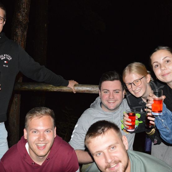 Fünf Personen posieren für ein Foto bei Nacht. Die beiden Frauen halten Getränke, und der Mann im grauen Hoodie lächelt. Ein Baumstamm und ein Holzzaun sind hinter ihnen.