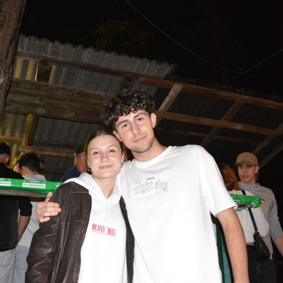 Ein Mann und eine Frau posieren nachts mit einem Holzgebäude im Hintergrund für ein Foto, jeweils in weißen und braunen Shirts.