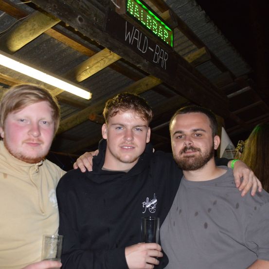 Drei Männer stehen zusammen, lächeln und halten Getränke in der Hand in einer Bar mit einem Schild, das Wald-Bar besagt.