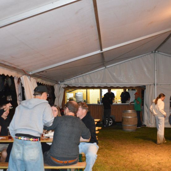 Ein Outdoor-Event unter einem Zelt, mehrere Personen sitzen an Tischen mit Getränken, während andere stehen. Eine Frau in Weiß geht auf das Zelt zu. Ein Fass steht neben dem Tresen, und ein Mann in einem grünen Shirt spricht mit einer anderen Person.