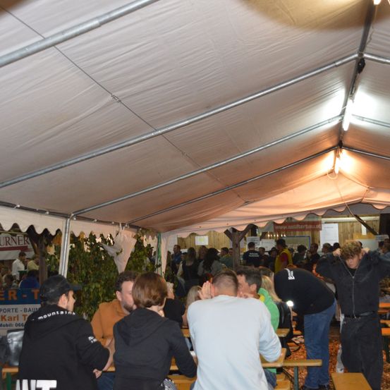 Eine große Gruppe von Menschen versammelt sich unter einem Zelt und isst an Tischen. Einige tragen Jacken. In der Ecke steht eine Pflanze.