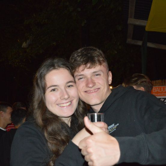 Ein junges Paar lächelt bei Nacht für die Kamera. Der Mann hält ein Glas und gibt einen Daumen hoch. Sie sind von Menschen umgeben, einige verschwommen, und im Hintergrund befindet sich ein gelbes Banner.