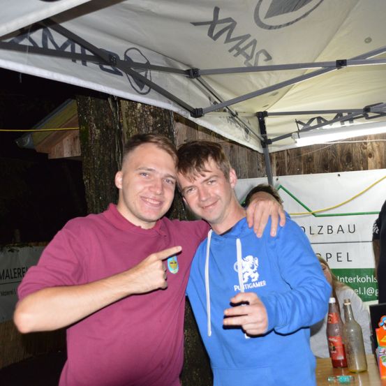 Zwei Männer posieren für ein Foto unter einem Zelt, einer trägt ein weinrotes Poloshirt und der andere eine blaue Kapuzenjacke. Sie lächeln und zeigen auf etwas, mit einem Banner und einem Baum im Hintergrund.