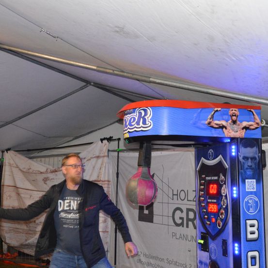 Ein Mann mit Brille und einer schwarzen Jacke mit einem weißen T-Shirt darunter spielt ein Box-Arcade-Spiel. Der Automat ist blau mit rotem Rand und zeigt eine digitale Punktzahl von 259 an.