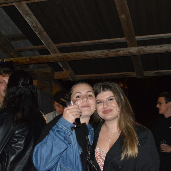 Zwei Frauen stehen eng beieinander und lächeln für ein Foto. Eine hält ein Getränk. Hinter ihnen unterhalten sich mehrere Leute. Die Umgebung ist schwach beleuchtet, mit einer Holzdecke.