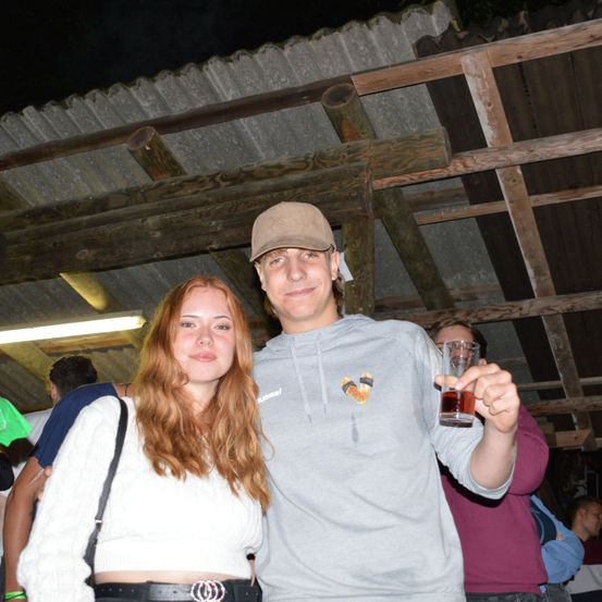 Ein junger Mann und eine junge Frau stehen lächelnd unter einem hölzernen Unterstand. Der Mann hält ein Glas Bier in der Hand und sie sind von anderen Menschen umgeben. Der Unterstand hat hölzerne Balken und ein Metalldach.