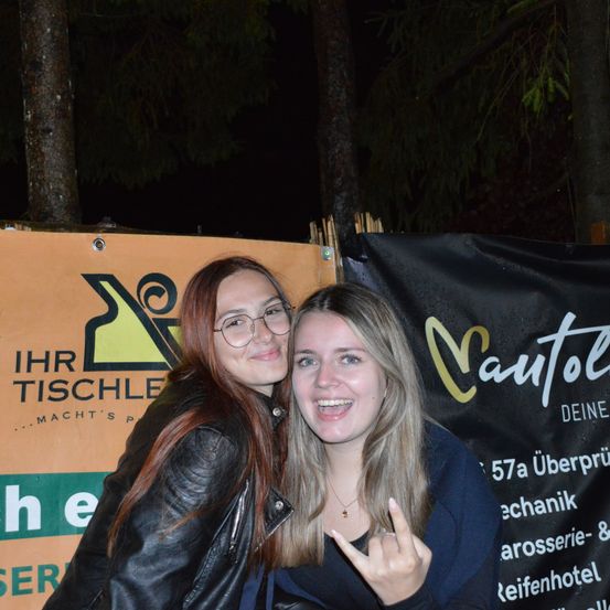 Zwei Frauen stehen eng beieinander und posieren für ein Foto bei Nacht. Die Frau auf der linken Seite trägt eine schwarze Lederjacke und eine Brille. Die Frau auf der rechten Seite macht ein Rockzeichen. Hinter ihnen sind Banner mit deutschem Text zu sehen.