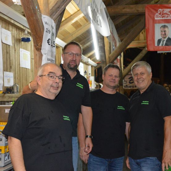 Vier Männer in schwarzen T-Shirts mit grünem Text vorne stehen in einem Raum mit Holzwänden und Holzbalken. Ein rotes Banner mit dem Foto eines Mannes hängt über ihnen.