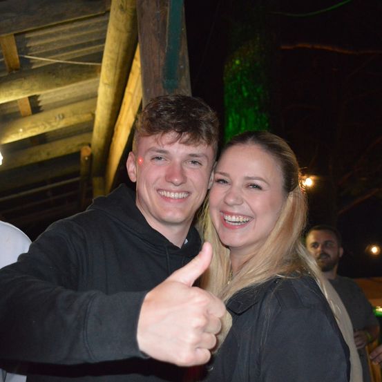 Ein junger Mann und eine junge Frau stehen eng beieinander, lächeln und posieren für ein Foto bei Nacht. Der Mann gibt einen Daumen hoch. Ein Mann im Hintergrund hält ein Getränk. Die Umgebung ist eine rustikale Holzstruktur mit einem grünen Licht.