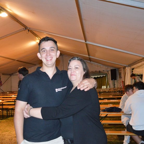 Ein Mann und eine Frau umarmen sich in einem Zelt, während andere Leute auf Bänken im Hintergrund sitzen.