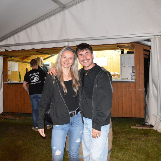 Zwei Personen stehen vor einem Essenszelt und lächeln. Ein Mann und eine Frau posieren eng zusammen. Dahinter steht ein Mann an einer Theke.