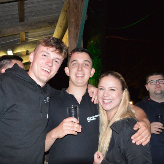 Drei junge Erwachsene, zwei Männer und eine Frau, lächeln und posieren für ein Foto bei Nacht. Sie halten Gläser in der Hand und scheinen sich auf einer gesellschaftlichen Veranstaltung zu befinden.