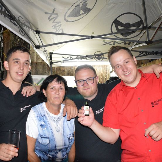 Vier Personen stehen unter einem Zelt mit Logo, zwei Männer in schwarzen und einer in rotem Shirt, und eine Frau in einer Jeansweste, posieren für ein Foto, wobei ein Mann eine kleine Flasche hält.