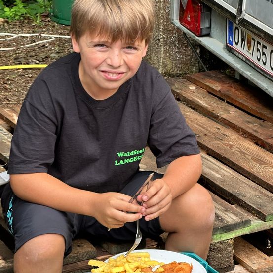 Ein Junge sitzt auf einer Holzbank, hält eine Gabel und ein Messer in der Hand und isst eine Mahlzeit. Er trägt ein schwarzes T-Shirt mit der Aufschrift 'Waldfest Langeco'. Hinter ihm steht ein Fahrzeug mit dem Kennzeichen 'OW 755' auf dem Boden geparkt.