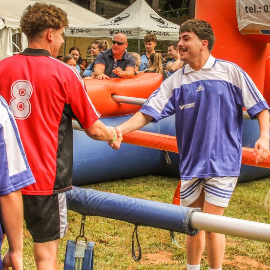 Zwei Jungen in Fußballtrikots schütteln sich die Hände vor einer orangefarbenen aufblasbaren Struktur. Andere Leute stehen und sitzen um sie herum. Der Junge links trägt ein rotes Trikot mit der Nummer 8.