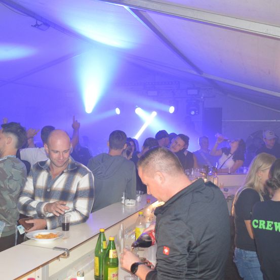 Eine Gruppe von Menschen in einer Bar. Der Tresen ist mit lila Lichtern beleuchtet. Einige Leute trinken und tanzen. Ein Barkeeper bereitet Getränke zu.