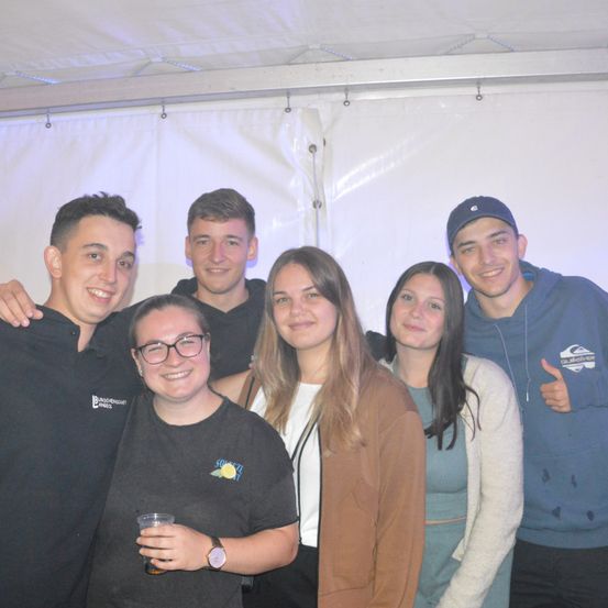 Eine Gruppe junger Erwachsener posiert für ein Foto unter einem weißen Zelt. Drei Männer und drei Frauen, alle lächelnd, wobei einer der Männer ein Getränk in der Hand hält.