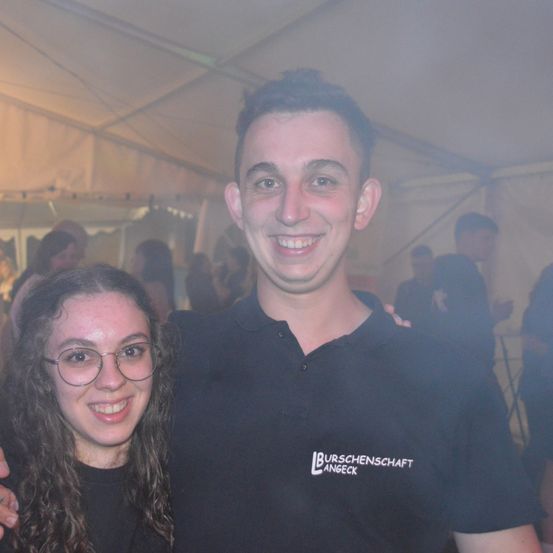 Ein Mann und eine Frau lächeln und posieren für ein Foto in einem Zelt. Der Mann hat seinen Arm um die Schulter der Frau gelegt. Der Hintergrund zeigt verschwommene Personen.