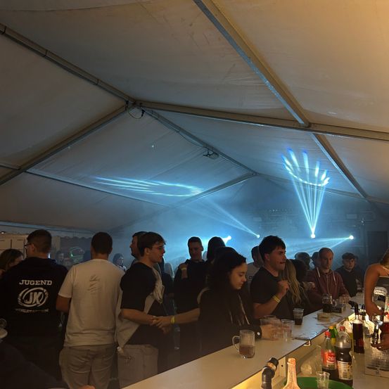 Eine Gruppe von Menschen steht um eine Bar in einem Zelt herum, beleuchtet von blauen Lichtern. Einige halten Getränke, während andere sich unterhalten.