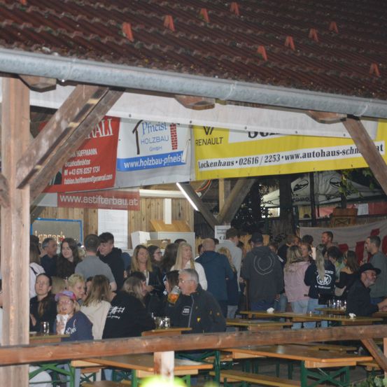 Ein überfülltes Bierzelt mit Menschen, die um Holztische und -bänke versammelt sind. Werbung für Holzbau Pfneisl und Renault ist auf dem Dach zu sehen.