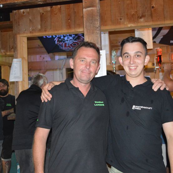 Zwei Männer in schwarzen Poloshirts mit Logos posieren für ein Foto in einer Bar. Hinter ihnen stehen andere Leute und eine Holzwand mit Postern.