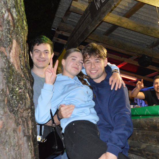 Drei Freunde posieren nachts für ein Foto, zwei Männer und eine Frau. Die Frau sitzt auf der Schulter eines Mannes, während ein anderer Mann sich an einen Baumstamm lehnt.