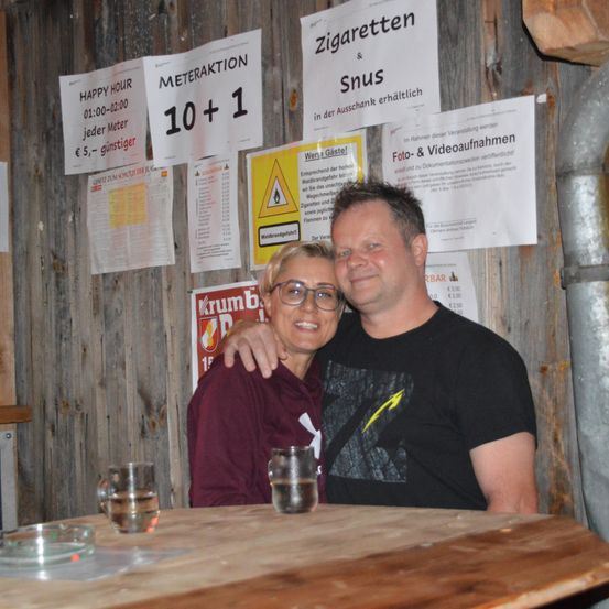 Ein Mann und eine Frau lächeln und posieren für ein Foto an einem Tisch. Hinter ihnen hängen mehrere Poster an einer Holzwand. Ein Poster sagt 'Meteraktion 10+1', ein anderes 'Happy Hour 01:00-02:00'. Gläser mit Wasser stehen auf dem Tisch.