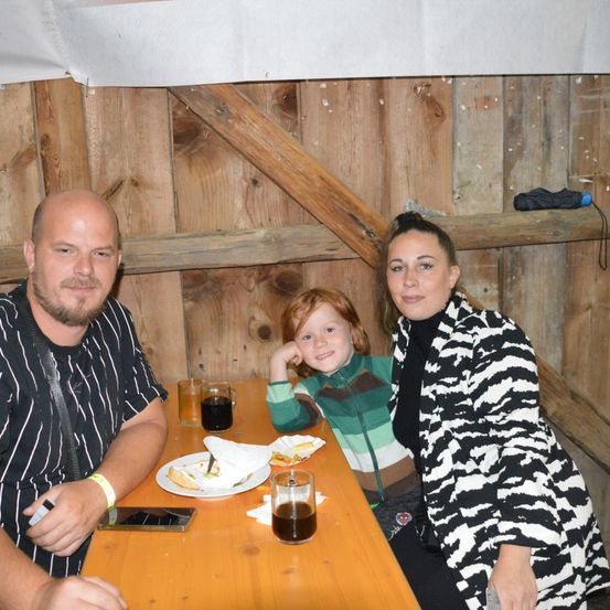 Eine dreiköpfige Familie sitzt an einem Holztisch. Ein Mann hält ein Handy, eine Frau lächelt und ein Kind stützt seinen Kopf auf die Hand. Teller, Servietten und Getränke stehen auf dem Tisch. Hinter ihnen ist eine Holzwand zu sehen.