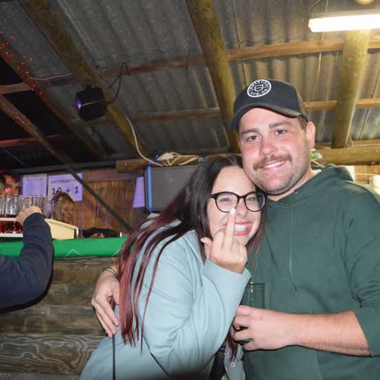 Ein Mann und eine Frau stehen lächelnd vor der Kamera. Die Frau hat rote Haare und Brille und hält ihren Finger hoch. Der Mann trägt einen grünen Hoodie und eine schwarze Baseballkappe. Hinter ihnen steht ein Schild mit der Aufschrift 'Meterakt 11'.