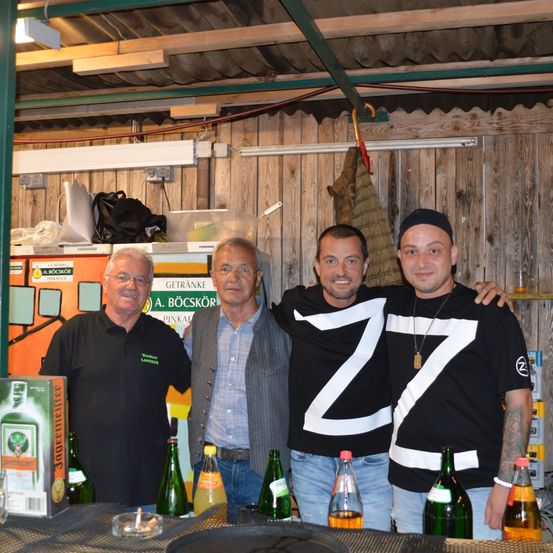 Drei Männer stehen in einem Raum mit Holzwänden und einer Bar. Der Mann links trägt ein schwarzes Shirt, der mittlere Mann einen grauen Anzug und der rechte Mann ein schwarzes Shirt mit weißen Buchstaben. Flaschen und eine Schachtel mit der Aufschrift Jaegermeister stehen auf der Theke.