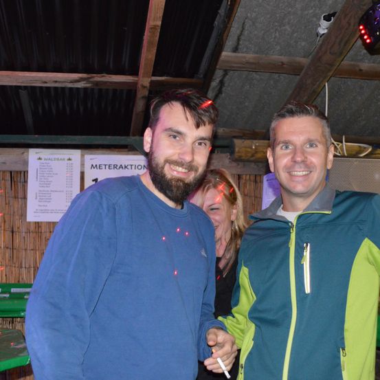 Zwei Männer lächeln und posieren für ein Foto mit einer Frau hinter ihnen in einer ungezwungenen Umgebung mit Schildern und Lichtern an der Wand.