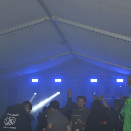 Eine Gruppe von Menschen tanzt in einem Club unter blauem Licht, einige tragen Brillen und Armbänder. Es gibt zwei Männer mit erhobenen Händen, einer hält ein Telefon und der andere ein Glas.