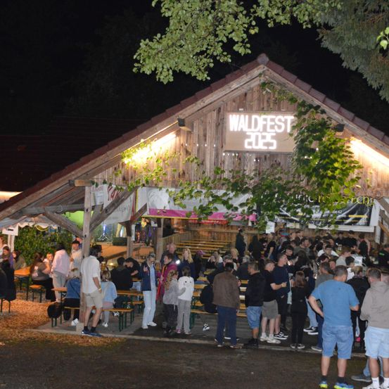Eine Nachtszene bei Waldfest 2023, Menschen versammeln sich um eine kleine Außenbar, einige sitzen auf Bänken, andere stehen, beleuchtet von warmen Lichtern.