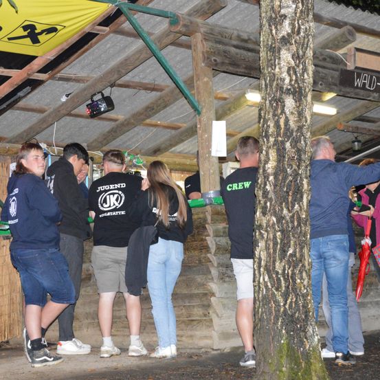 Eine Gruppe von Menschen in Freizeitkleidung steht in einer rustikalen Umgebung mit Holzwänden und einem gelben Banner. Sie scheinen in der Nähe eines Baumes in einer Schlange zu warten.