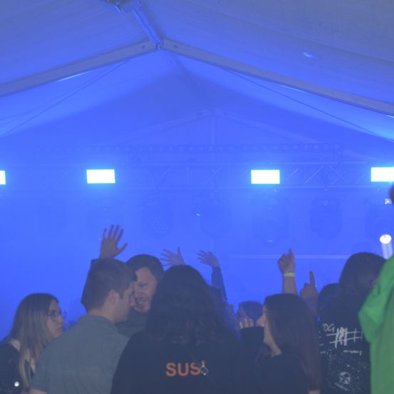 Eine Gruppe von Menschen befindet sich in einem Zelt mit blauen Lichtern. Einige Menschen heben ihre Hände. Die Person vorne trägt ein schwarzes Shirt mit weißen Buchstaben.