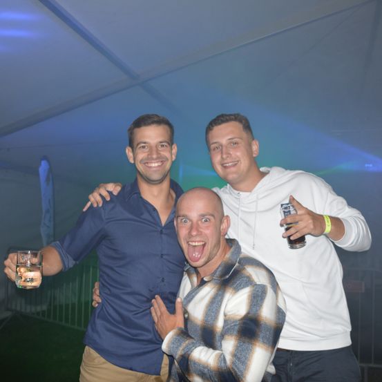 Drei Männer posieren für ein Foto unter einem Zelt mit blauen Lichtern. Ein Mann hält ein Glas Wasser, ein anderer ein Bier und der Mann in der Mitte hat seine Zunge herausgestreckt.