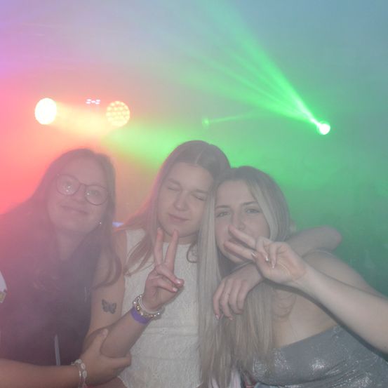 Drei Frauen posieren für ein Foto in einem Club mit bunten Lichtern, eine zeigt ein Friedenszeichen, eine andere eine Handgeste und die dritte lächelt.