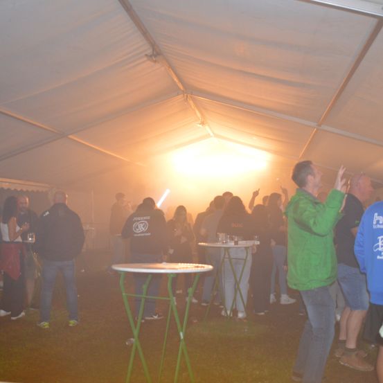 Eine Menschenmenge versammelt sich unter einem großen Zelt. Einige stehen, andere sitzen. Ein Mann in einer grünen Jacke hebt die Hände. Es gibt Tische mit Getränken und ein helles Licht im Hintergrund.