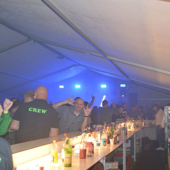 Eine Gruppe von Menschen versammelt sich in einem Zelt mit blauer Beleuchtung. Ein Mann in einem schwarzen T-Shirt mit grünem Text und ein anderer in einem grauen Hemd stehen neben einer Bartheke mit verschiedenen Getränken.