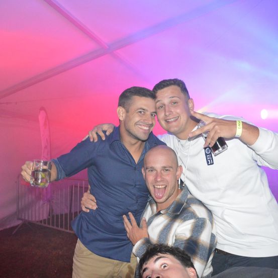 Drei Männer lächeln und posieren für ein Foto bei einer Party. Ein Mann hält ein Getränk, und ein anderer hat ein gelbes Armband. Sie stehen unter farbiger Beleuchtung.