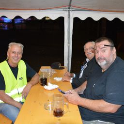 Drei Männer sitzen bei Nacht unter einem Zelt. Einer trägt eine neongelbe Weste, ein anderer trägt eine Brille und der dritte hält ein Handy. Sie haben einen Tisch mit Getränken und Servietten.