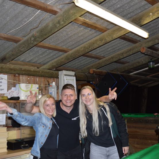 Drei Personen posieren für ein Foto in einem Raum mit Holzwänden und einer Metalldecke. Ein Mann in der Mitte macht Handzeichen. Eine Frau auf der linken Seite trägt eine Jeansjacke und macht ein Friedenszeichen. Eine weitere Frau auf der rechten Seite lächelt, während sie den Mann umarmt.